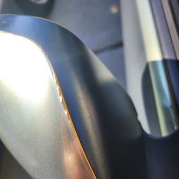 Wrapros - The image features a close-up of a vehicle's side mirror, which has a metallic silver finish with a slight shimmer. The mirror's edge is outlined with a thin orange line, and the surrounding trim is black. Part of the vehicle's window and door are visible in the background, along with a blurred view of a parked car and pavement. The lighting suggests it is daytime with sunlight reflecting off the mirror's surface. - Vehicle Wraps Near Me - Montclair, California