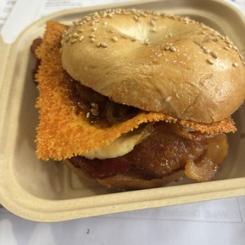 Hound and Mare - The Melrose - A breakfast sandwich featuring a bagel with sesame seeds, bacon, fried fish fillet, and caramelized onions, served in a takeout container. - Cafes Near Me - Ithaca, New York
