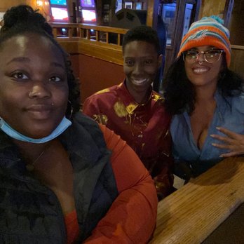 Gabby's Bar - three women sitting at a bar - Four individuals, three women and one man, are seated at a wooden table in a bar or restaurant setting. The woman on the left wears a black jacket, a blue face mask hanging below her chin, and hoop earrings. The man in the middle wears a red shirt with yellow floral patterns. The woman on the right wears glasses, a colorful beanie, a blue shirt, and has her hand on her chest. The background features dim lighting and wooden decor. - Sports Bars Near Me - Columbus, Ohio