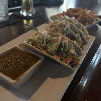 Trophy - Tacos - A rectangular white plate holds three tacos filled with shredded meat, chopped vegetables, and shredded cheese, garnished with shredded cheese on top. A small square dish of salsa is placed beside the tacos. In the background, there are additional plates of food and a glass of beer on a dark wooden bar counter, with a blurred bar setting visible. - Sports Bars Near Me - Chandler, Arizona