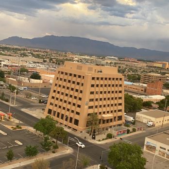 DoubleTree by Hilton Hotel Albuquerque - View from 14th floor - The image captures a city view from a high vantage point, featuring a large beige office building with a clock on its facade. Surrounding the building are parking lots with several parked cars, trees lining the streets, and a network of roads. In the background, a mountain range is visible under a cloudy sky, indicating an urban area near natural landscape. - Hotels Near Me - Albuquerque, New Mexico