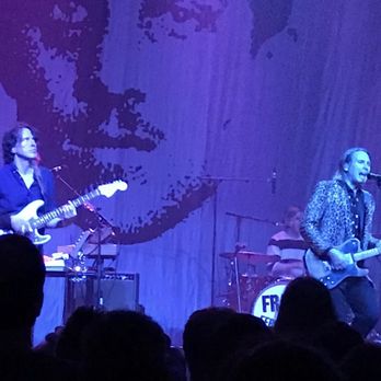 Rams Head Live - A band performs live on stage with multiple musicians, including a guitarist, drummer, and vocalist, each playing their instruments and singing into microphones. The stage has a large portrait backdrop, and the audience is visible in the foreground. The venue appears to be a spacious concert hall with professional lighting and sound equipment. - Music Venues Near Me - Baltimore, Maryland