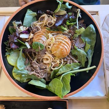Gom Ramen - We totally took a bite before taking the picture ‍ - A bowl of ramen contains a mixture of fresh salad greens, thin noodles, and two eggs topped with sauce. The dish is presented in a black bowl with a wooden rim, placed on a wooden tray. The background shows a person wearing a green and blue plaid shirt, and part of a table with a small container of sauce or dressing. - Noodles Near Me - El Paso, Texas