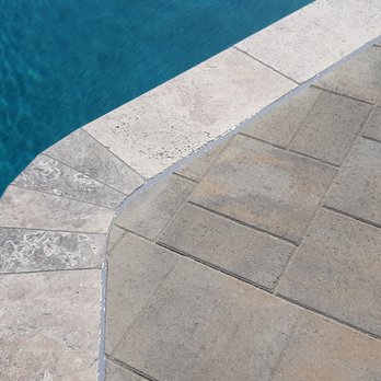 Baccaro Pools - A photograph captures the edge of a swimming pool with blue water on the left and a tiled concrete deck on the right. The pool's curved border is lined with light-colored stone tiles, contrasting with the rectangular paving stones of the deck. The image emphasizes the clean, geometric design of the pool area, focusing on the intersection of water and hardscape surfaces. - Pool Cleaners Near Me - Walnut, California