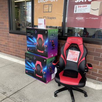 Staples - A red and black gaming chair with adjustable armrests and a high backrest is positioned on a sidewalk outside a store. Two boxed gaming chairs, matching the design of the assembled chair, are stacked beside it. The store window behind displays signs for Amazon returns and drop-off services, with visible pricing and promotional information. - Shipping Centers Near Me - Whitehall, Pennsylvania