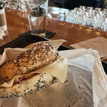 Yorgo's Bageldashery - Ham and Cheese bagel - A sesame seed bagel sandwich with cheese and meat is held in a hand over a tray at a cafe, with a glass of water and bar counter in the background. - Bagels Near Me - Norfolk, Virginia