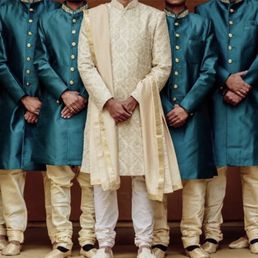 Men in matching teal kurtas with gold buttons and cream-colored churidars, with one man in the center wearing a cream sherwani, pose against a plain background.