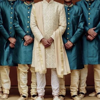 Sari Palace - Men in matching teal kurtas with gold buttons and cream-colored churidars, with one man in the center wearing a cream sherwani, pose against a plain background. - Traditional Clothing Near Me - Berkeley, California
