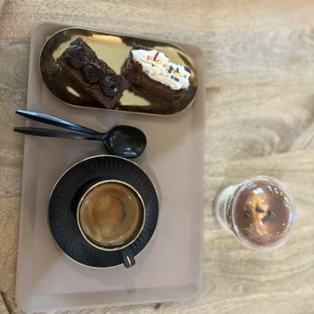 LiBre Dessert Cafe - A beige tray contains two slices of chocolate cake with frosting, one decorated with whipped cream and colorful sprinkles, a black cup of coffee with a saucer, and a cold beverage in a plastic cup with ice and a straw, all placed on a wooden table. - Coffee & Tea Near Me - Hamburg, New Jersey