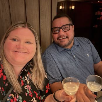 Silver Lyan - A woman with long blonde hair and a man with short dark hair and glasses sit at a table in a restaurant with wooden paneling and warm lighting. They are smiling and holding cocktail glasses with light-colored drinks. The background features dim lighting and dark interior decor, indicating an indoor dining setting. - Cocktail Bars Near Me - Washington, DC, District of Columbia