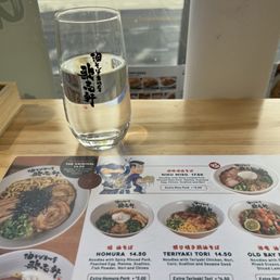 A clear glass of water with Japanese characters printed on it is placed on a wooden table near a window. The menu on the table displays different ramen dishes, including options with spicy minced pork, teriyaki chicken, and other toppings, with prices listed. The background shows a street scene with bicycles and buildings visible through the window.