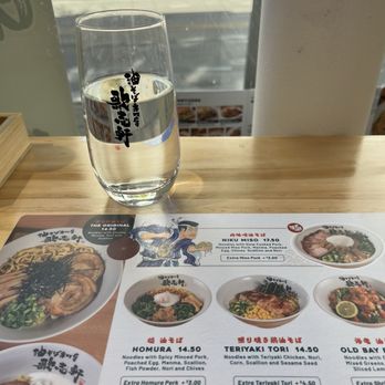 Kajiken - A clear glass of water with Japanese characters printed on it is placed on a wooden table near a window. The menu on the table displays different ramen dishes, including options with spicy minced pork, teriyaki chicken, and other toppings, with prices listed. The background shows a street scene with bicycles and buildings visible through the window. - Ramen Near Me - Champaign, Illinois