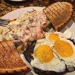 Black Diamond Steak - A breakfast plate includes two sunny-side-up eggs with runny yolks, two slices of toasted bread, and a serving of a creamy dish topped with a sauce and seasonings. The plate is placed on a speckled brown countertop, and the food appears freshly prepared.