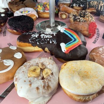 Voodoo Doughnut - Multiple donuts with different toppings and decorations are displayed on a pink tray at Voodoo Doughnut in Austin, TX. The donuts include chocolate, sprinkles, fruit, and themed designs, with some featuring icing, sprinkles, and colorful decorations. The tray has labels for banana cream pie and mango tart, indicating flavor options. - Donuts Near Me - Austin, Texas