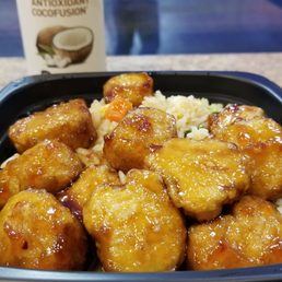 Orange Chicken - The image shows a black plastic takeout container filled with orange chicken coated in a glossy sauce, placed on a table. The dish is accompanied by a portion of white rice with small vegetable pieces. In the background, there is a cup with a coconut flavoring label, indicating a beverage or flavoring option. The setting appears to be a casual dining or takeout environment at Panda Express in Chicago.
