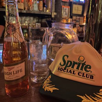 Pubkey - Pleb (Miller Hight Life, Campari, and Sweet Vermouth) - The image shows a bar counter with a Miller High Life beer bottle, a glass filled with water and ice, a glass jar containing cash, and a Sprite Social Club cap with a black brim and yellow embroidery. The background features shelves with various bottles of alcohol, framed pictures, and neon signs, indicating a lively bar environment. - Pubs Near Me - Washington, DC, District of Columbia