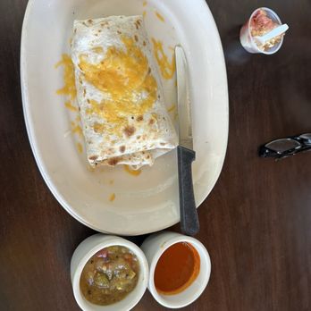 Flying Star Café - Breakfast burrito - A breakfast burrito with melted cheese on top is served on a white tray, accompanied by two small bowls of salsa and a side of yogurt with a spoon. - Bakeries Near Me - Albuquerque, New Mexico