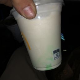 a hand holding a cup of milkshake - A hand with a visible thumb and fingernail holds a McDonald's McCafé vanilla milkshake in a clear plastic cup with a lid. The milkshake appears creamy and light-colored. The background shows part of a vehicle interior, including a seat and some objects, indicating the photo was taken inside a car.