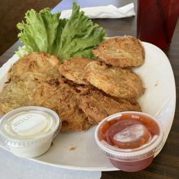 Fried green tomatoes - A white plate holds several fried green tomatoes with a crispy coating, garnished with a few leaves of lettuce. Two small plastic containers of dipping sauces, one creamy and one red, are placed on the plate. The plate is on a dark wooden table, with a red drink in a tall glass and a napkin in the background.