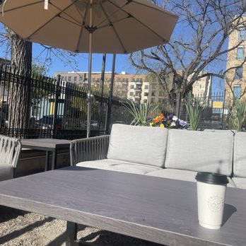 Schoolyard Cafe - The image shows an outdoor seating area with a rectangular table, a coffee cup, a sofa with light-colored cushions, and a large umbrella providing shade. There are additional chairs and a small side table nearby. The background includes a black metal fence, trees, colorful flowers, and multi-story buildings, indicating an urban environment. The scene is well-lit with clear skies, suggesting daytime. - Ice Cream & Frozen Yogurt Near Me - Denver, Colorado