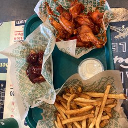 a tray of chicken wings and french fries - A green plastic tray contains fried chicken wings, French fries, and glazed chicken wings, each lined with paper printed with green text. A small cup of dipping sauce is also on the tray. The tray is placed on a table with a paper placemat and a partially visible beverage bottle.