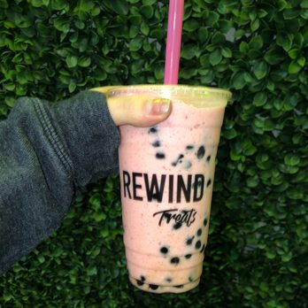Rewind - Butterfly Strawberry Lemonade caffeine free dairy free with boba - A hand, wearing a dark gray sleeve, holds a large clear plastic cup filled with pink strawberry lemonade bubble tea containing black tapioca pearls. The cup has a pink straw and features the 'REWIND Treats' logo printed on it. The background consists of dense green leaves, providing a natural setting for the beverage display. - Bubble Tea Near Me - Chandler, Arizona