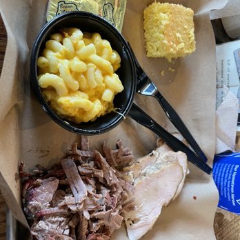 Mission BBQ - Brisket and Turkey with Mac n cheese - A meal tray contains sliced smoked meat, macaroni and cheese in a black cup, a piece of cornbread, and two small condiment packets. The food is served on a paper-lined tray, with a pair of black sunglasses placed on the side. The setting appears to be casual, likely at a barbecue restaurant. - Barbeque Near Me - Springfield, Illinois