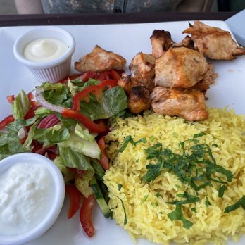 Thyme - Chicken kebab - A square white plate holds a serving of grilled chicken pieces, yellow rice topped with chopped herbs, a side salad with lettuce, tomato slices, and red onion, and two small cups of sauces—one creamy and one possibly mayonnaise or yogurt-based. The meal is set on a table, with part of a person's torso visible in the background. - New American Near Me - Roslyn, New York