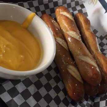 Outpost - Bread pretzels & molten cheese - Three baked pretzel breadsticks with slashed tops and coarse salt sit on checkered paper, accompanied by a cup of melted cheese for dipping. - American Near Me - Anoka, Minnesota