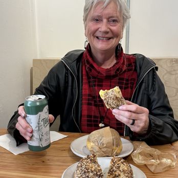 Sunday Bagels - My Cheeky Monkey..:) - A woman with short gray hair, wearing a red plaid shirt and black jacket, is sitting at a wooden table with bagels and a can of drink, smiling. - Bagels Near Me - Albuquerque, New Mexico