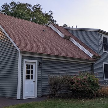 Fiji Construction - Rear of house before gutters were hung. - A two-story residential house with gray vinyl siding, a brown gable roof, and several windows. It has a garage with a white door and a side door near some bushes. - Roofing Near Me - Cary, Illinois