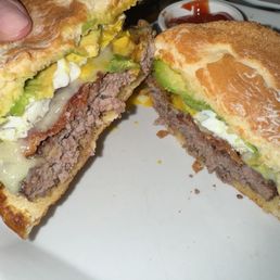 Breakfast Burger - A burger with a beef patty, avocado slices, egg, and melted cheese, cut in half on a white plate, with a small bowl of ketchup in the background.