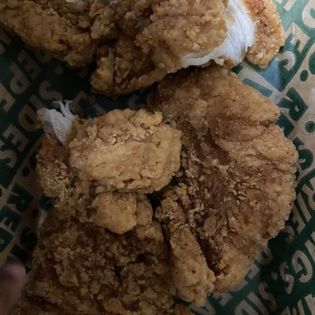 Wingstop - Burnt chicken tenders - The image displays several pieces of fried chicken with a crispy, textured coating. The chicken pieces vary in size and shape, with some torn open to show white meat inside. They are placed on branded paper with green and white text, likely from a fast-food or fried chicken restaurant. The focus is on the fried chicken's exterior, which appears crunchy and well-cooked. - Chicken Wings Near Me - Woodbridge, New Jersey