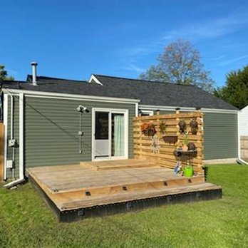 Thermalcraft - The image shows a backyard with a wooden deck attached to a green house. The deck has a small step down to a grassy lawn. A wooden privacy wall with a vertical garden of potted plants is on one side of the deck. The background includes a blue sky, trees, and neighboring houses. - Windows Installation Near Me - Villa Park, Illinois