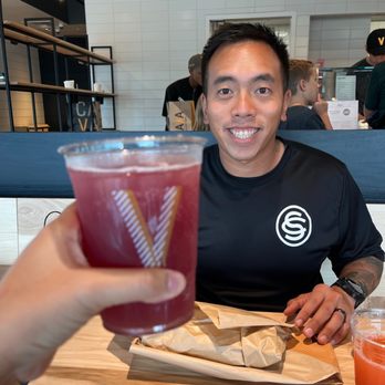 CAVA - A man is seated at a restaurant table, smiling at the camera, with a drink held in the foreground. The background shows a modern restaurant interior with other patrons. - Mediterranean Near Me - Georgetown, Texas