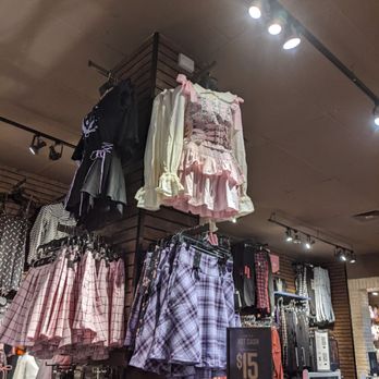 Hot Topic - The image shows a retail clothing store with various skirts and dresses on display. Prominently, there are pink and purple plaid skirts hanging on a rack, along with a pink Lolita-style dress with ruffles, bows, and lace details. The store has wooden paneling on the walls and ceiling-mounted lighting fixtures illuminating the merchandise. A sign indicates a $15 sale price for some items. - Accessories Near Me - Glendale, Arizona