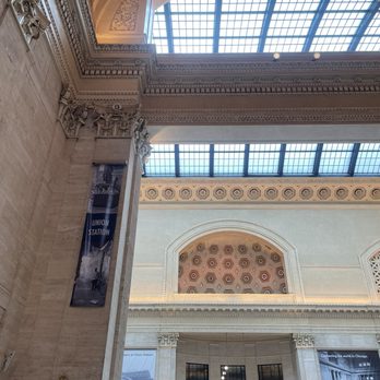 Amtrak - The photo captures the interior of Chicago Union Station, highlighting its classical architectural elements such as decorative moldings, columns, and detailed cornices. The ceiling includes large, arched skylights with a grid pattern, allowing natural light to illuminate the space. There are banners and signs indicating services like Amtrak, customer service, and ticketing, with a clock visible near the center. The overall scene emphasizes the historic and grand design of the station's interior. - Trains Near Me - Chicago, Illinois