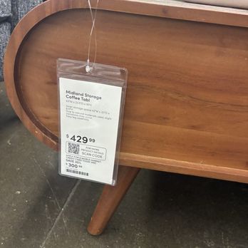 World Market - A wooden coffee table with a rounded edge and tapered legs is shown in a furniture store. A price tag is hanging from the corner of the table, displaying the price of $429.99 and product details. The background includes other furniture items and textiles, indicating an indoor retail environment. - Home Decor Near Me - Albuquerque, New Mexico
