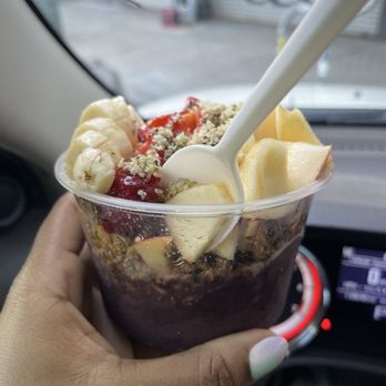 Main Squeeze - Alpha bowl - A hand with a manicured nail holds a clear plastic cup containing an acai bowl topped with banana slices, strawberries, and other fruits, with a white spoon inserted. The background shows the interior of a vehicle, including part of the dashboard and windshield, with an outdoor scene visible through the windshield. - Juice Bars & Smoothies Near Me - Oakland, California