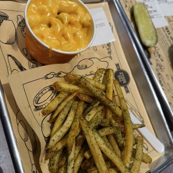 The Melt - GARLIC PARMESAN FRIES  MINI MAC - The image shows a tray with a paper liner from The Melt restaurant, featuring seasoned French fries with herbs, a cup of cheese macaroni and cheese, and a pickle spear. The tray is placed on a metal surface, with additional paper liners and a pickle on a separate tray in the background. - Burgers Near Me - Tempe, Arizona