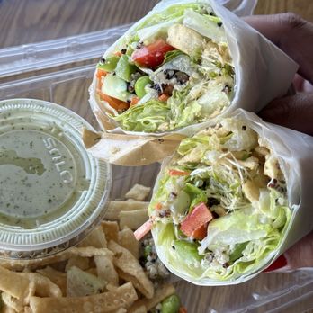 Toss’d - Avocado Wrap - Two rice paper wraps filled with fresh vegetables such as lettuce, tomatoes, and possibly cucumbers, are held in hand over a plastic tray. A side of crispy chips and a small cup of green herb dipping sauce are also on the tray. The wraps are partially unwrapped, revealing the colorful vegetable filling inside. - Wraps Near Me - Temecula, California