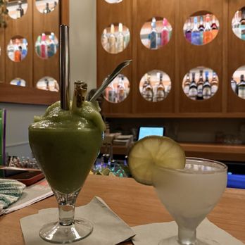 Daydream - Two cocktails are positioned on a wooden bar counter, one is a green frozen drink served in a tall glass with a straw and spoon, and the other is a clear beverage garnished with a lemon slice in a short glass. The background features a wooden liquor display with circular cutouts showcasing various bottles, and a computer monitor is visible behind the bar. The setting appears to be a bar or restaurant interior. - Cocktail Bars Near Me - Albuquerque, New Mexico