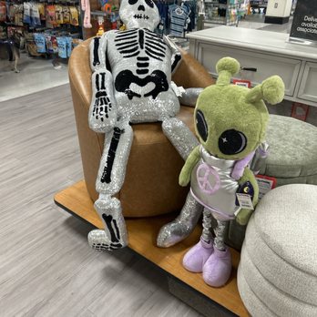 TJ Maxx - Hurry for those great Halloween finds. - A plush skeleton toy with a shiny, sequined surface and black details is sitting on a store bench, leaning back against a brown cushion. Next to it, a green alien plush toy with large black eyes, antennae, and a peace sign on its chest stands upright. The store background features shelves with various toys, clothing, and accessories, indicating a retail environment, likely a department or toy store. - Department Stores Near Me - Pasadena, California