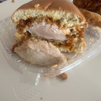 Filly Luv - Crazy N' Luv - Hot Sandwich - A partially eaten chicken sandwich with a bun, chicken breast, and toppings is placed in a clear plastic container on a white surface. The sandwich shows the inside ingredients, including the chicken and toppings, with some crumbs and pieces of chicken outside the sandwich. In the background, there is another sandwich with a knife on top, partially visible. - Chicken Shop Near Me - Brooklyn, New York