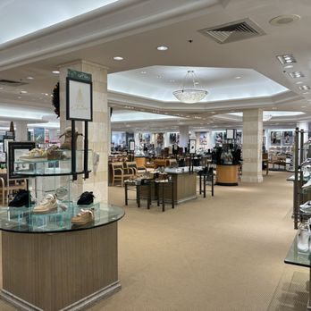 Dillard's - The image depicts the interior of a Dillard's department store, with shoe displays on glass-topped tables and shelves along the walls. Seating areas with chairs are visible, along with Christmas decorations and signage. The ceiling has recessed lighting and a central chandelier, and the space is well-lit with a neutral color scheme and beige carpeting. - Department Stores Near Me - Overland Park, Kansas