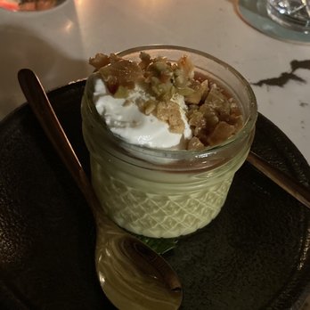 Papi - Flan - A dessert of flan served in a small glass jar, topped with whipped cream and chopped nuts, placed on a black plate with a spoon. - Cocktail Bars Near Me - Portland, Maine
