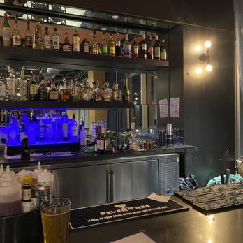 Blu Violet - Bar - The image shows a bar area with multiple shelves of liquor bottles, including spirits and liqueurs, arranged above a stainless steel counter. The bar is equipped with various bar tools, glasses, and a blue-lit beverage dispenser. A drink in a glass is placed on the counter in the foreground. The background includes a mirror reflecting part of the bar and some ambient lighting fixtures, with curtains on the right side of the scene. - Cocktail Bars Near Me - Providence, Rhode Island