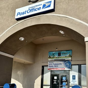US Post Office - The photo depicts the exterior of a U.S. Post Office building featuring a prominent sign with the United States Postal Service logo and text. There are mail collection boxes positioned outside near the entrance. Above the door, there are signs related to passport applications and travel photos. The building has an arched entryway and a beige facade with minimal decorative elements. - Post Offices Near Me - Murrieta, California