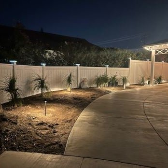 Smith Landscape - Delicate, Ponytail Palms exquisitely contrast against the white fencing. - The image shows a landscaped outdoor area at night, featuring small plants and shrubs illuminated by ground lighting. A white privacy fence runs along the background, and a curved concrete pathway is visible in the foreground. The scene appears to be part of a residential or commercial property with maintained landscaping. - Masonry/Concrete Near Me - Upland, California