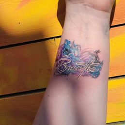 Tattoo of cooled toned smoke with the word Breathe in black with white accents written in a script font over it.